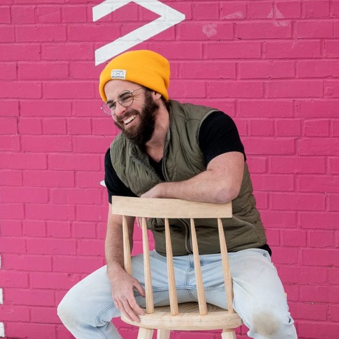 Bo Morin sits backwards on a wooden chair in front of a pink wall, wearing a yellow hat and green vest